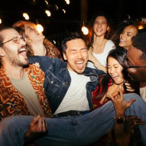 A multicultural group of friends are lifting their male tipsy friend at the rooftop night outdoor party.