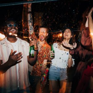 A cheerful group of multiracial friends is celebrating and having fun with confetti at the open-air summer rooftop nightclub. They are having a blast, dancing and drinking beer.